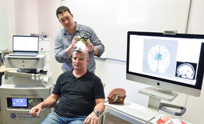 A man sits on a chair as a second man stands behind him using an ultrasound device on his head.
